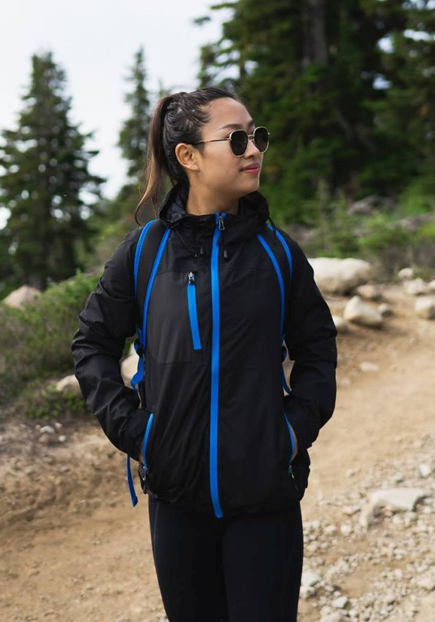 Woman in a black shell jacket with blue trim on a forest trail.