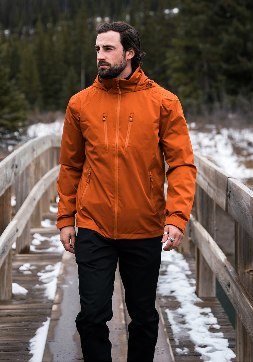 Man in an orange lightweight shell jacket on a snowy wooden footbridge.